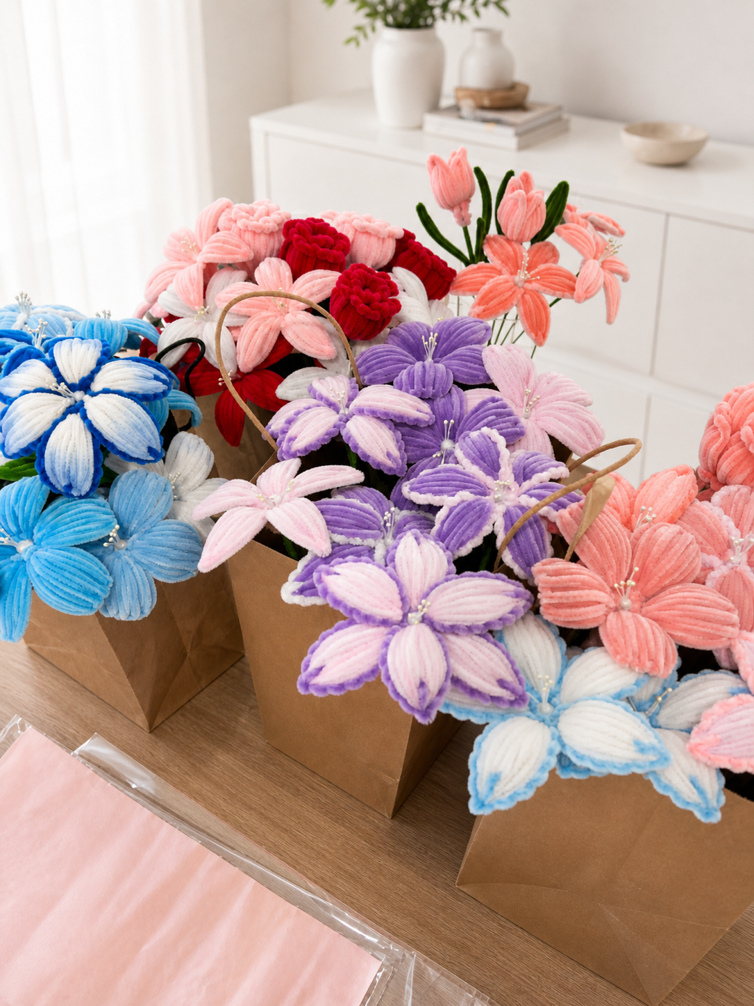 A row of finished pipe cleaner bouquets in soft blue, red, pink, purple and peach, packed into kraft paper carry bags ready to be delivered